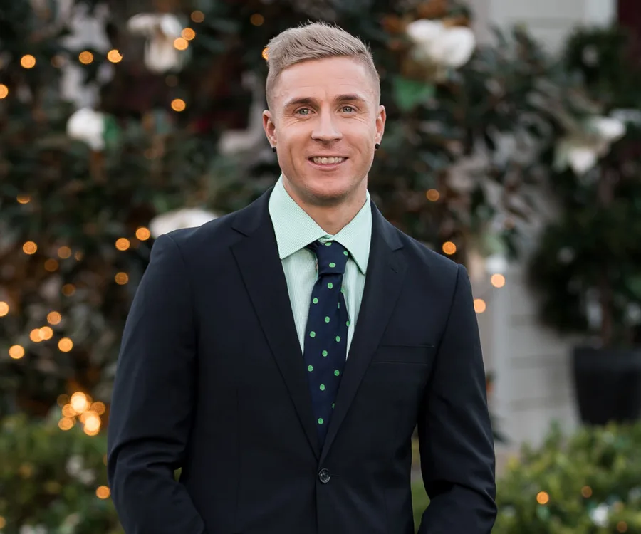Man in a suit with a light green shirt and polka-dotted tie, smiling against a blurred, festive floral background.