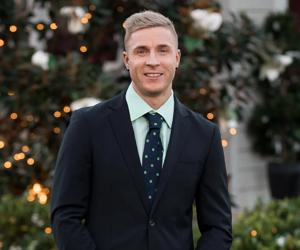 Man in a suit with a light green shirt and polka-dotted tie, smiling against a blurred, festive floral background.