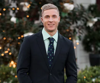 Man in a suit with a light green shirt and polka-dotted tie, smiling against a blurred, festive floral background.