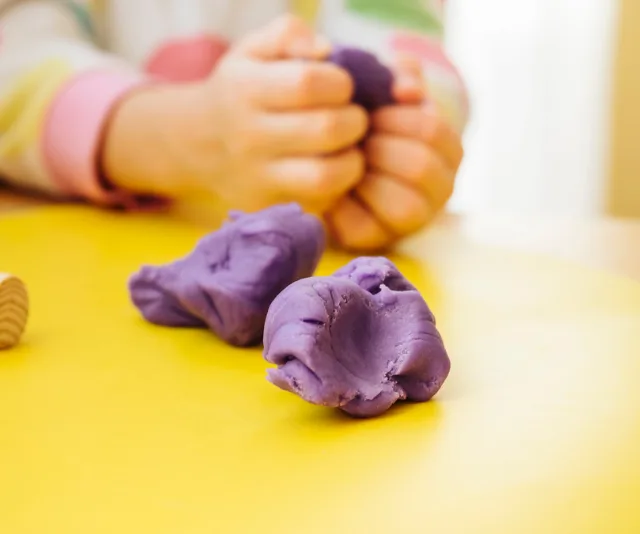 Child's hands molding purple playdough on a yellow table.