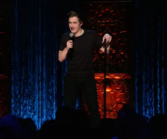 Comedian performs on stage holding a microphone, standing against a decorative wall and blue curtain.