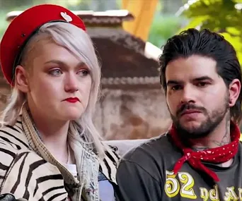 A couple sits closely, looking serious. The woman wears a red hat and zebra-patterned jacket; the man sports a red bandana.