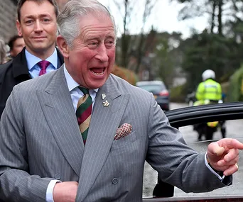 Prince Charles smiling and gesturing, wearing a striped tie and suit, with a man in a suit behind him by a car.