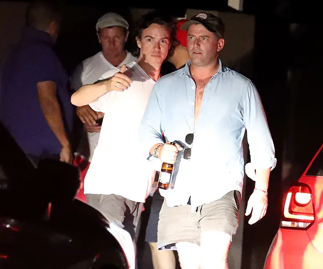 Group of people at night, two men in foreground; one holding a drink, wearing a cap; others in casual attire.