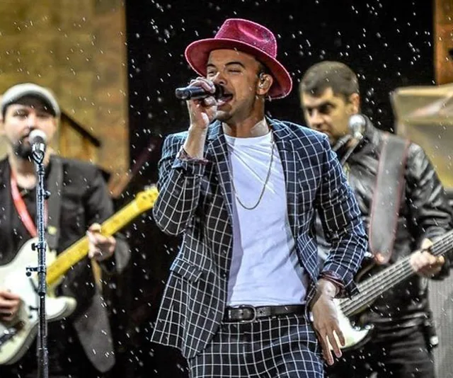 Singer in a patterned suit and red hat performs on stage with two guitarists in the background.