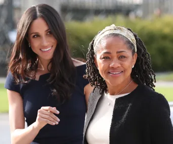 Two women smiling outdoors, one with long dark hair in a blue dress and the other with locs and a headband in a black jacket.