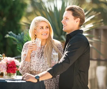 A couple smiling, holding drinks outdoors with flowers and greenery around them.