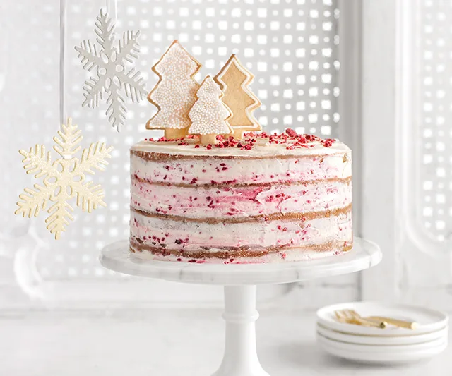 Festive cake with white and pink frosting, topped with tree-shaped cookies; snowflake decorations in the background.