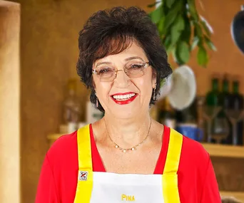 Pina smiling in a kitchen setting, wearing a red top and a yellow-trimmed apron.