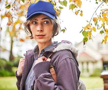 A person wearing a blue cap and a fur-lined jacket stands outdoors beside autumn leaves.