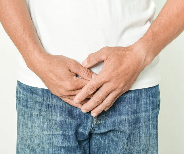 Man in jeans and white shirt holding hands near groin area, suggesting discomfort or health issue.
