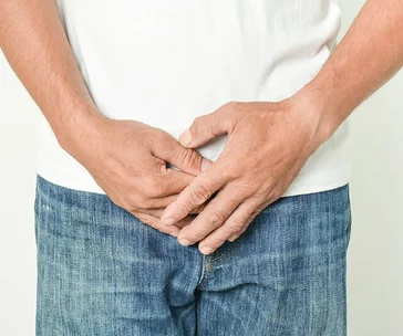 Man in jeans and white shirt holding hands near groin area, suggesting discomfort or health issue.