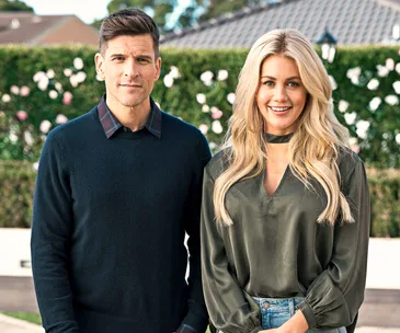 Man in dark sweater and woman in green blouse smiling, standing outdoors with a hedge and flowers in the background.
