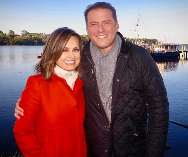 A man and woman smiling by a waterfront, with trees and boats in the background.