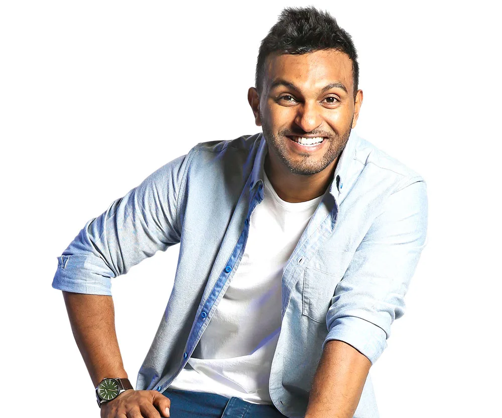 A person smiling, wearing a light blue shirt over a white t-shirt, leaning slightly forward against a white background.