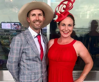 A couple dressed stylishly at the Melbourne Cup; man in a plaid suit and hat, woman in a red dress and matching fascinator.