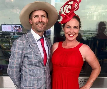 A couple dressed stylishly at the Melbourne Cup; man in a plaid suit and hat, woman in a red dress and matching fascinator.