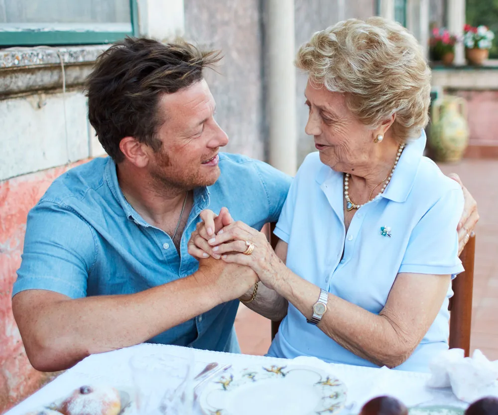 Jamie Oliver smiling and holding hands with an elderly woman at an outdoor table with plates and food items.