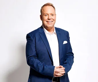 A man in a blue suit and white shirt smiling with hands clasped in front, against a plain white background.