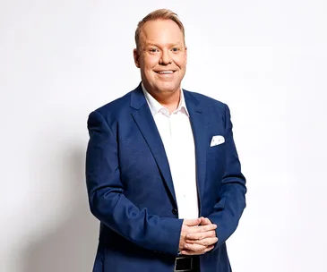 A man in a blue suit and white shirt smiling with hands clasped in front, against a plain white background.
