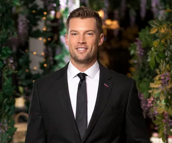 Man in a black suit with a black tie, smiling, standing amidst greenery and decorative lights.