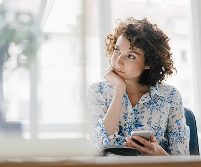 A woman in a floral blouse sits thoughtfully, holding a smartphone, looking out of a sunlit window.