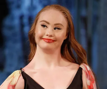 A young woman with long reddish-brown hair and floral sleeve top smiling on a runway.