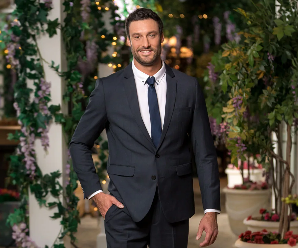 Man in a suit poses with greenery and lights in the background.