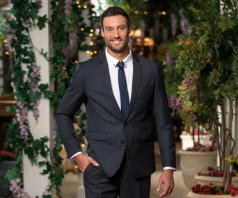 Man in a suit poses with greenery and lights in the background.