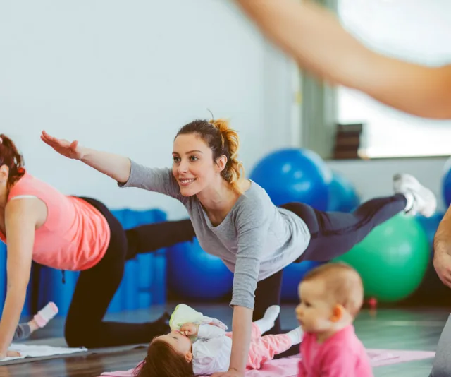 Women exercising in a class with babies, stretching on mats with exercise balls in the background.