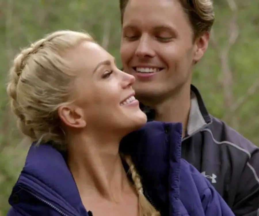 A couple shares an affectionate moment outdoors; woman with blonde braid, man in dark jacket smiling with eyes closed.