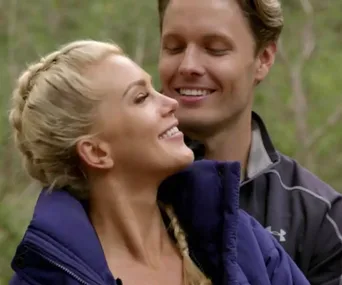 A couple shares an affectionate moment outdoors; woman with blonde braid, man in dark jacket smiling with eyes closed.