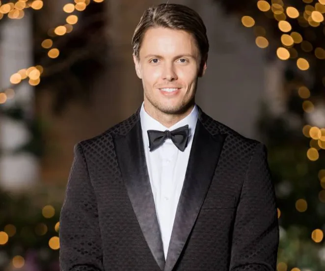 A man in a formal black tuxedo with a black bow tie, smiling, with blurred decorative lights in the background.