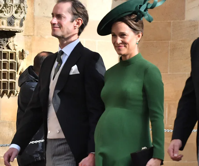 A couple dressed formally, with the woman wearing a green maternity dress and hat, smiling as they walk outdoors.