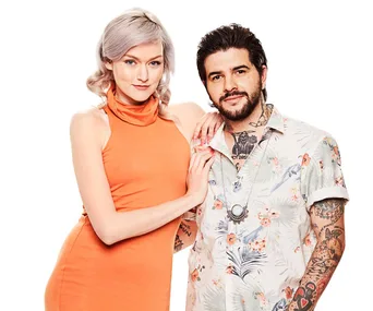 A couple poses together, the woman in an orange dress and the man in a floral shirt with tattoos, on a white background.