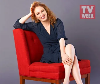A woman with curly red hair, wearing a navy dress, sits smiling on a red chair against a gray background with "TV WEEK" logo.