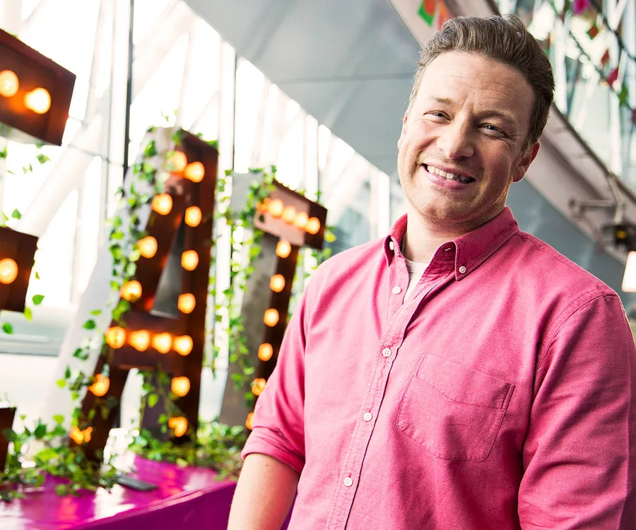 Man in pink shirt smiling, standing indoors with decorative illuminated letters in the background.