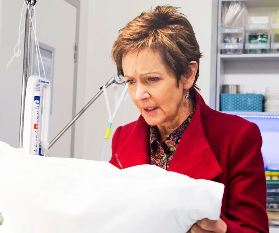 Susan in a red coat stands in a tense hospital scene, holding a white pillow, looking concerned.