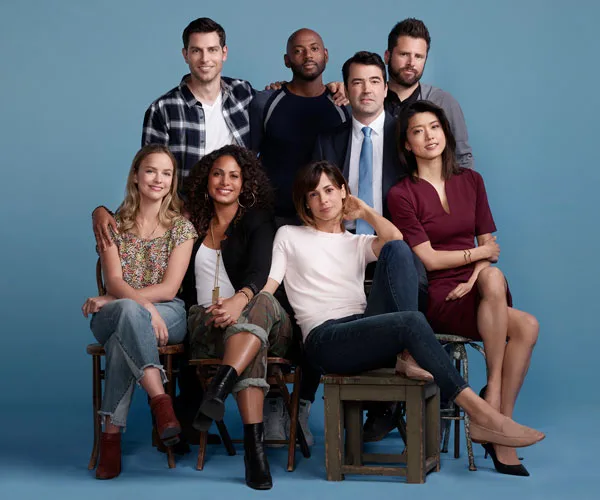 Cast of "A Million Little Things" seated and standing together against a blue background, smiling and posing for a group photo.