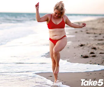 Woman in a red bikini joyfully walks along a beach, with the ocean in the background.