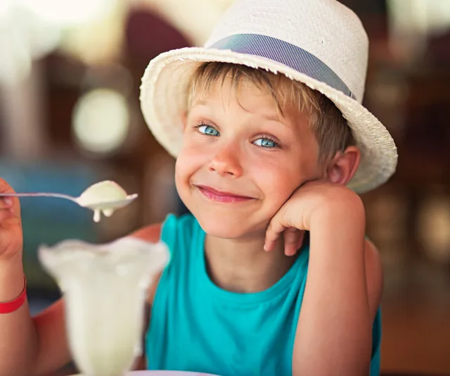 Young boy in a white hat, smiling, holding a spoon near a dessert glass in a casual setting.