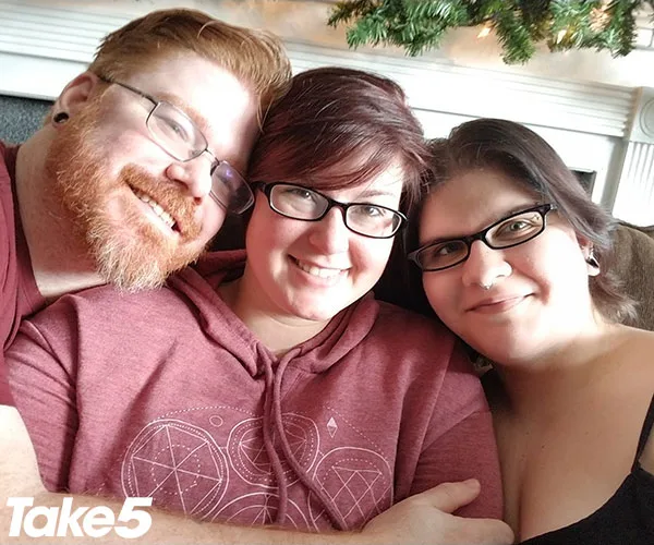 Three people smiling and sitting close together, wearing glasses, in a cozy indoor setting with greenery above.