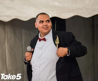 Man in a suit with a red bow tie holding a microphone, appears to be speaking or performing at an event.