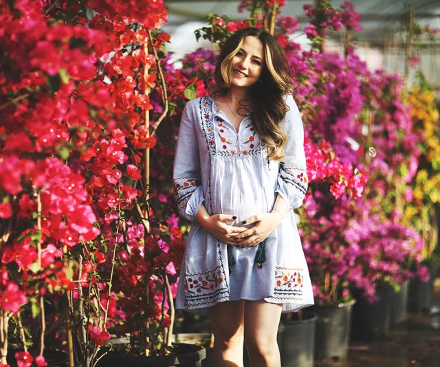 Pregnant woman in embroidered dress stands smiling among vibrant flowering plants.