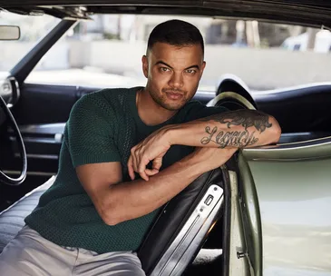 Guy Sebastian sitting in a car, wearing a green shirt, with a tattooed arm resting on the seat.