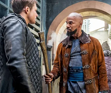 Two characters from "Robin Hood" (2018) face each other, one holding a wooden staff.