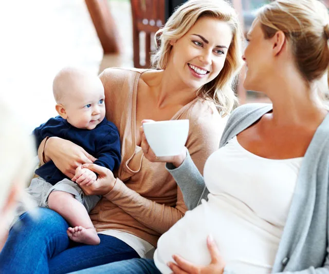 Pregnant woman with coffee cup chatting with woman holding baby, both smiling warmly indoors.