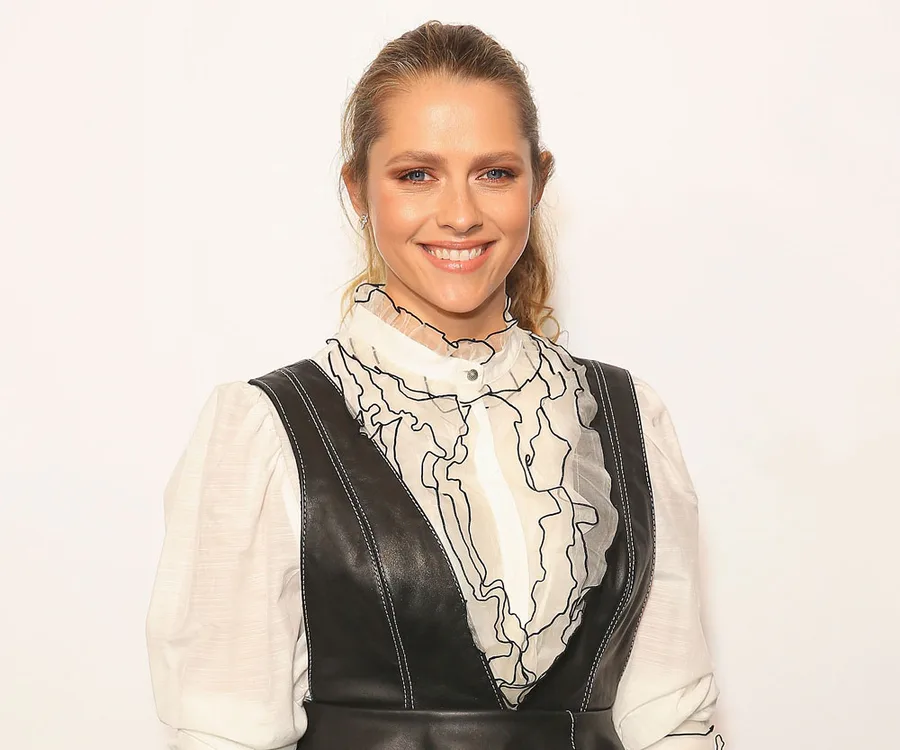 Teresa Palmer smiling in a ruffled white blouse with a black vest at a photoshoot.