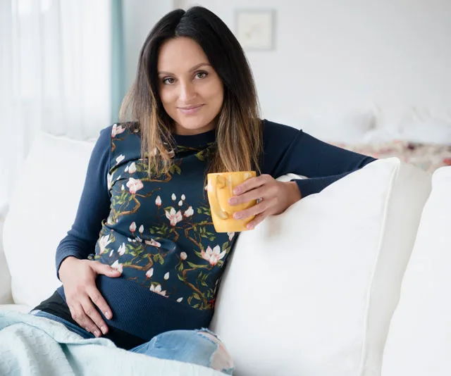 Pregnant woman relaxing on a couch, holding a yellow mug and gently touching her belly, wearing a floral-patterned top.