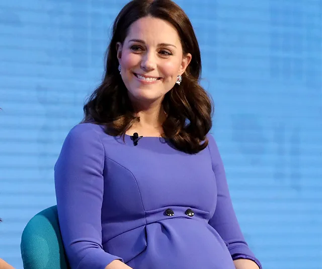 A pregnant woman in a purple dress smiles while seated.